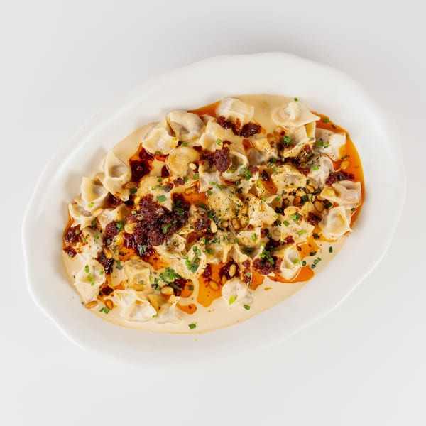 Plate of dumplings in creamy sauce with chili oil, crispy sausage, pine nuts, and chopped herbs.
