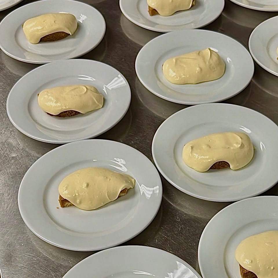 Multiple small pastries topped with thick pale frosting are arranged on white plates across a stainless-steel kitchen counter.