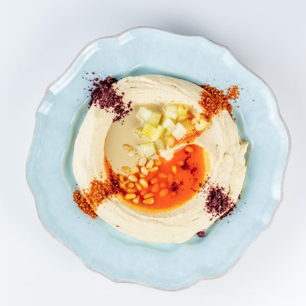 Overhead view of hummus topped with diced vegetables, pine nuts, chili oil, and colorful spices on a blue plate.