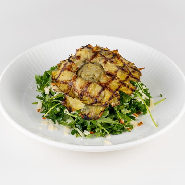 Grilled artichoke hearts served over a leafy green salad with shaved cheese and toasted nuts on a white plate.