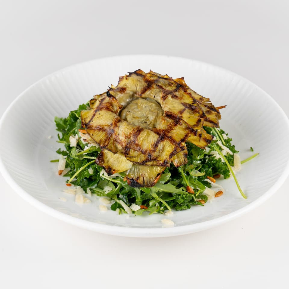 Grilled artichoke hearts served over a leafy green salad with shaved cheese and toasted nuts on a white plate.
