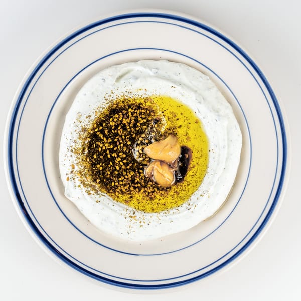 Creamy yogurt dip topped with olive oil, za’atar, sesame seeds, and roasted garlic on a blue-rimmed plate.