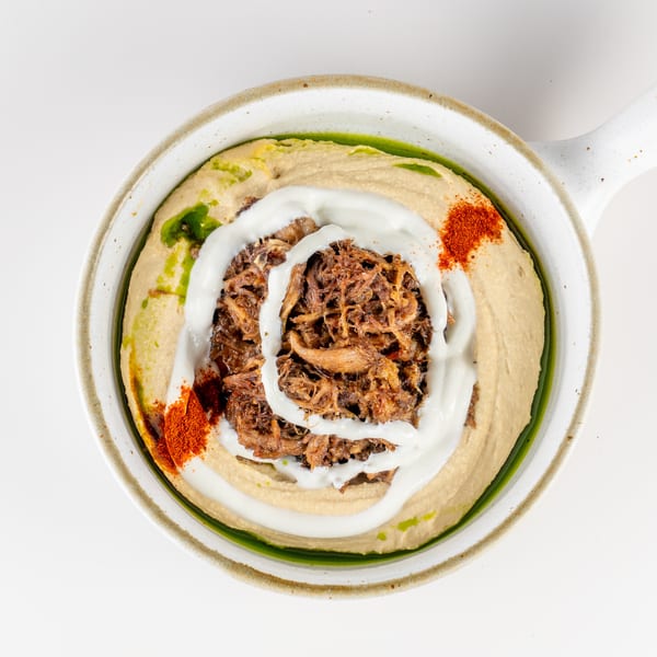 Bowl of hummus topped with shredded meat, yogurt sauce, olive oil, and red spices.