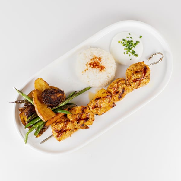 Grilled chicken skewer with white rice, roasted vegetables, and herb-topped dipping sauce on a white plate.