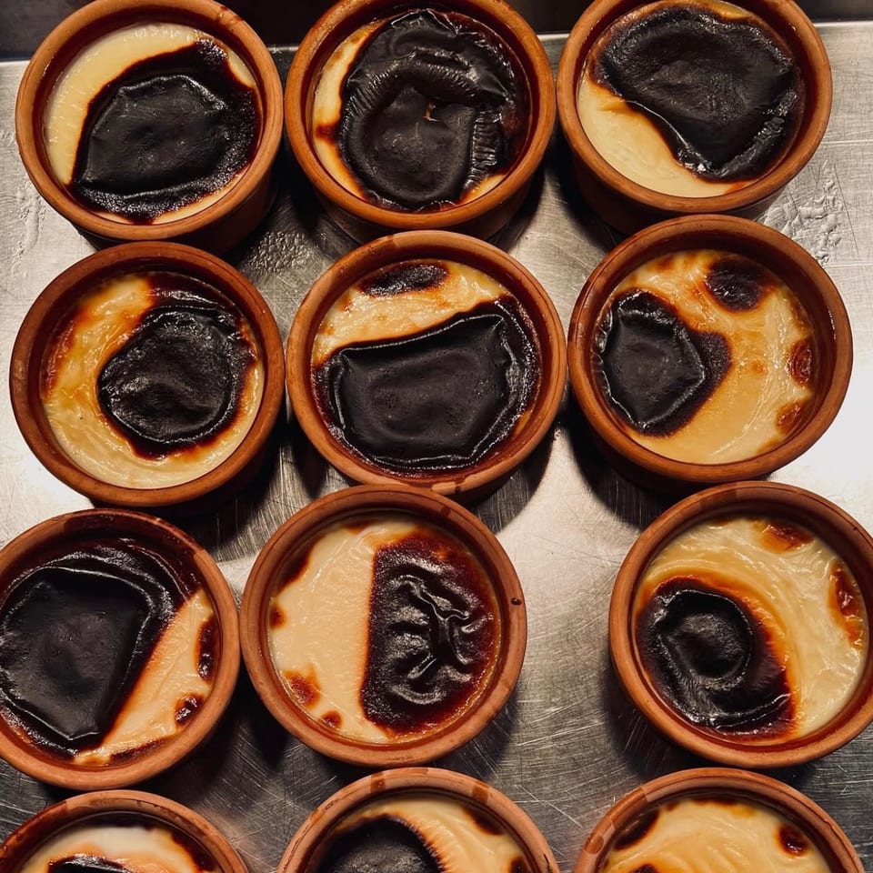 Overhead view of custard tarts in terracotta dishes, topped with dramatically dark, caramelized surfaces on a metal tray.