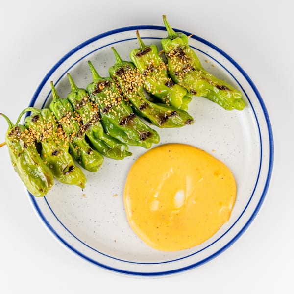 Grilled green shishito peppers topped with sesame seeds, served beside a pool of orange dipping sauce on a blue-rimmed plate.