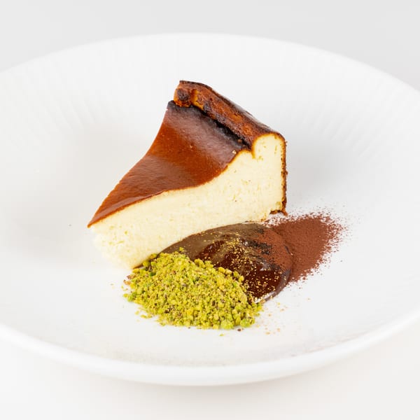 Slice of Basque cheesecake with chocolate sauce, crushed pistachios, and cocoa powder on a white plate.