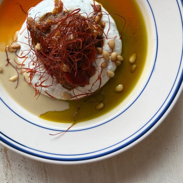 Creamy yogurt dish topped with roasted vegetables, pine nuts, chili threads, tahini, and olive oil on a blue-rimmed plate.