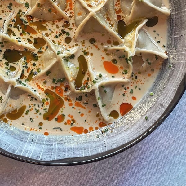 Overhead view of folded dumplings in creamy sauce, topped with chopped chives and colorful chili oil, served on a glass plate.