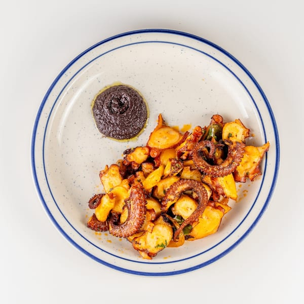 Grilled octopus and roasted potatoes with vegetables, served alongside dark dipping sauce on a blue-rimmed plate.