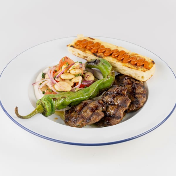 Grilled meat kebabs with roasted green peppers, bean salad, and toasted bread topped with red pepper sauce on a plate.