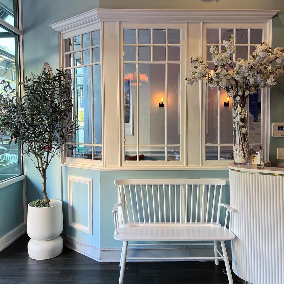 Elegant restaurant entryway with a white bench, potted tree, floral arrangement, candlelit windows, and blue paneled walls.