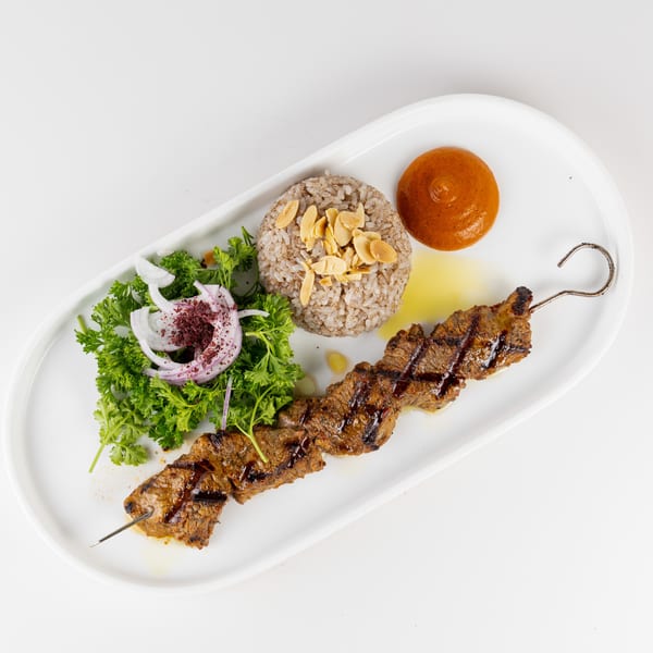 Grilled meat skewer served with almond-topped rice, parsley and onion salad, and a round bread roll on a white plate.