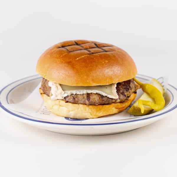 Grilled cheeseburger with creamy sauce on a toasted bun, served with pickles and sliced peppers on a plate.