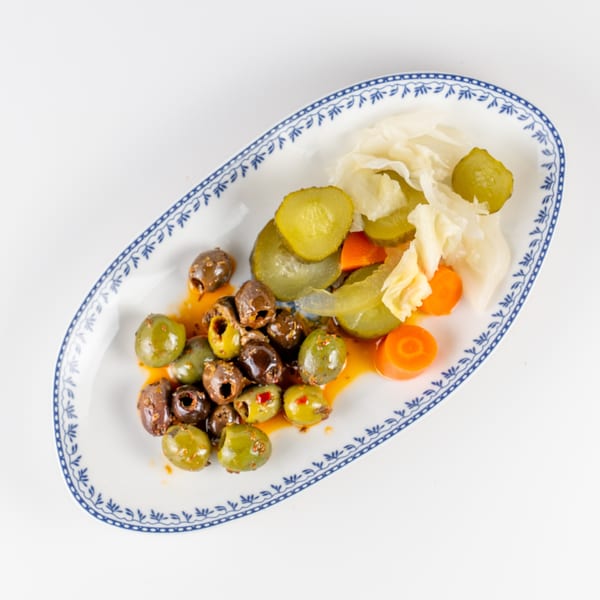 Mixed green and black olives, pickles, carrots, and cabbage served on a blue-and-white patterned platter.