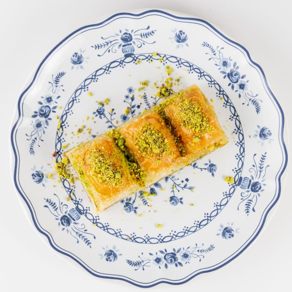 Three golden baklava pieces topped with crushed pistachios on a decorative blue-and-white floral plate.