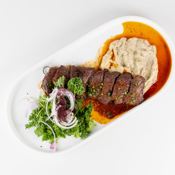 Grilled beef skewers served with creamy hummus, spiced butter sauce, parsley, and sliced red onions on a white plate.