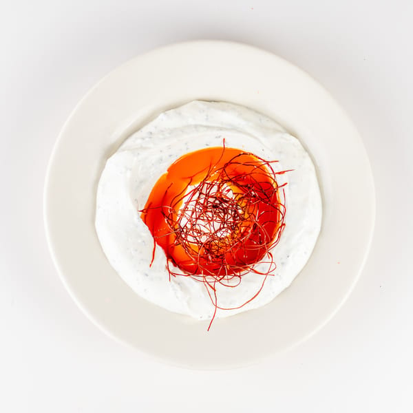 Overhead view of creamy white sauce surrounding a vibrant orange-red center, topped with delicate red spice threads on a white plate.