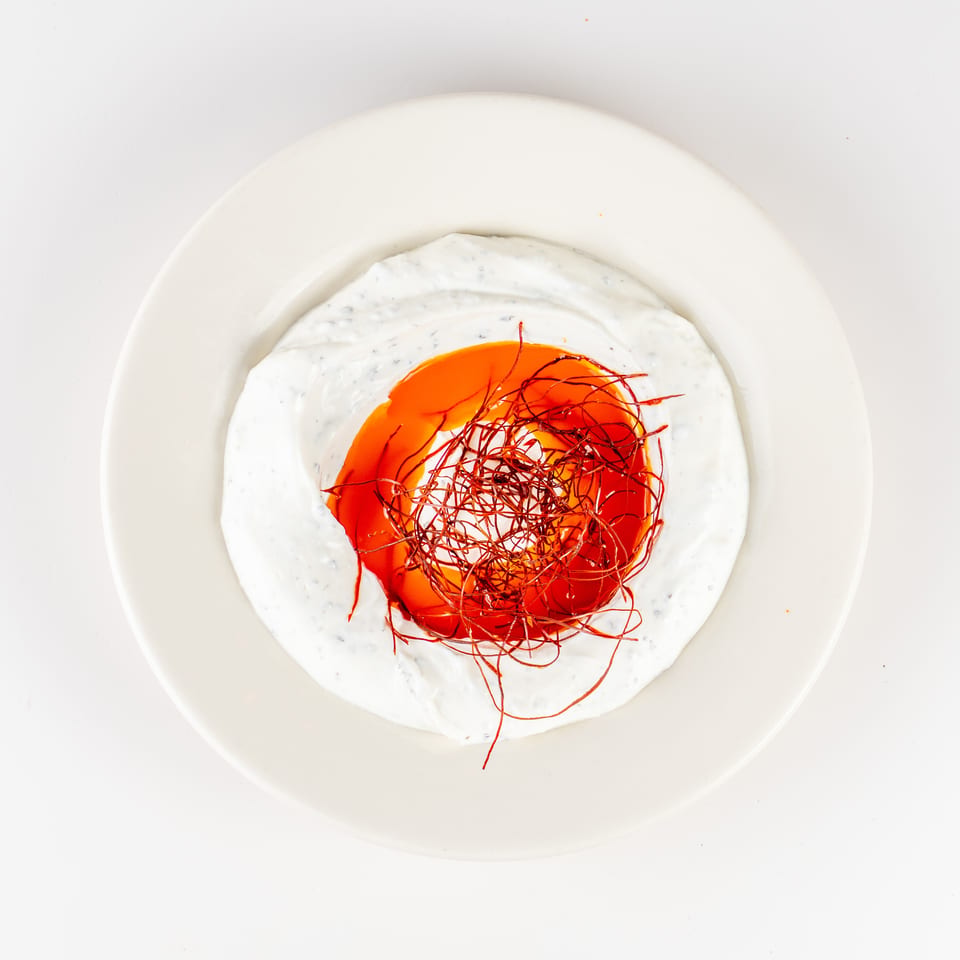 Overhead view of creamy white sauce surrounding a vibrant orange-red center, topped with delicate red spice threads on a white plate.